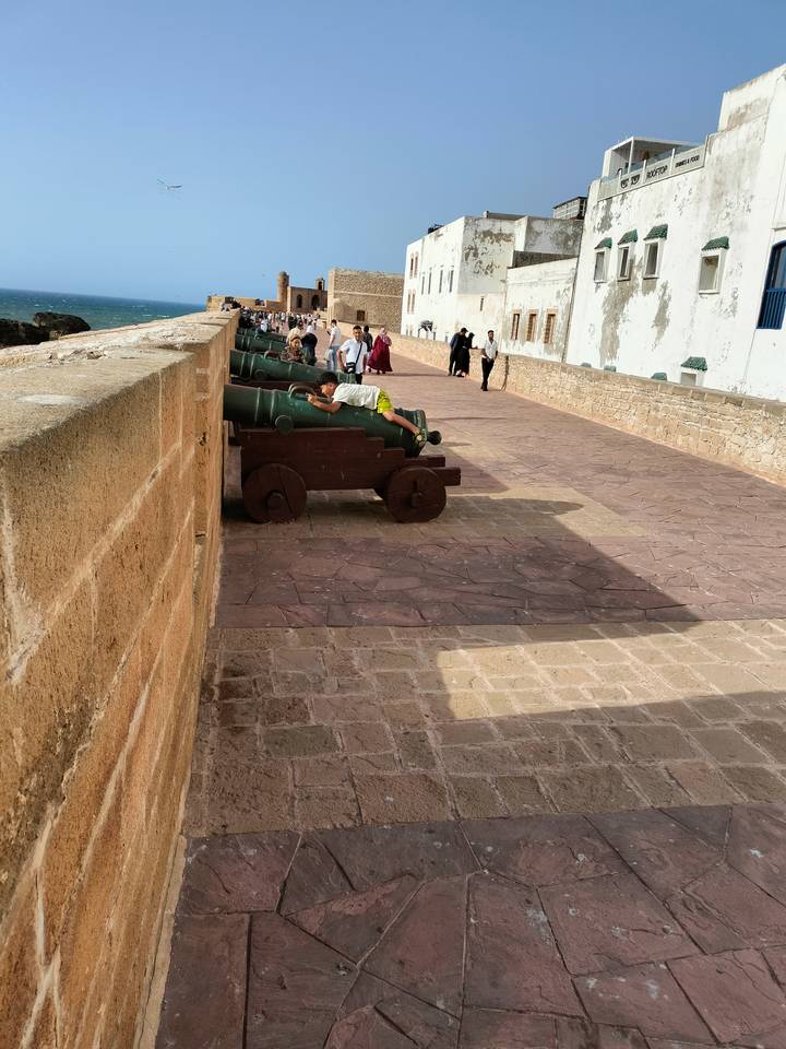 Stone rampart with historic cannons where tourists stroll and a child climbs onto one cannon.