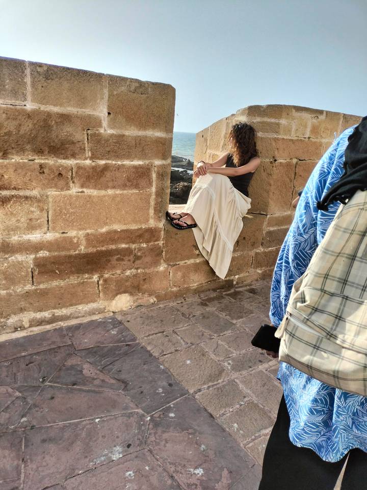 Partial shot of a woman sitting on a stone sill with ocean in background, framed by another passer-by’s clothing.