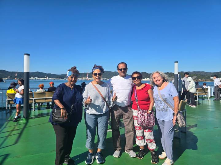 Five mature travellers pose on the open deck of a ferry with blue sea and wooded hills in the distance.