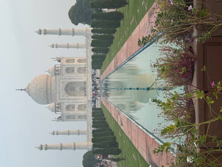 The Taj Mahal reflected in its central pool with rows of cypress trees and visitors in the distance.