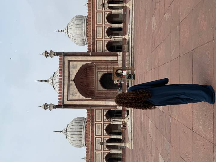 A woman in a long abaya stands before the grand red-sandstone arches of Jama Masjid in Delhi.