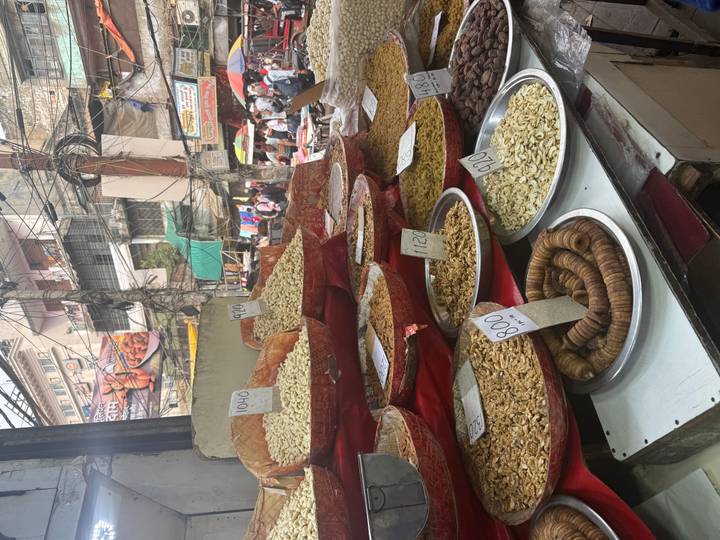 A bustling street-market stall displays mounds of nuts and dried fruit with handwritten price tags.
