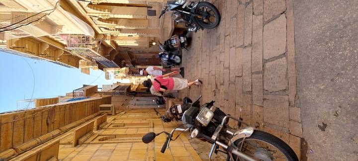 Narrow sandstone alley lined with motorbikes and tourists exploring in sunlight.