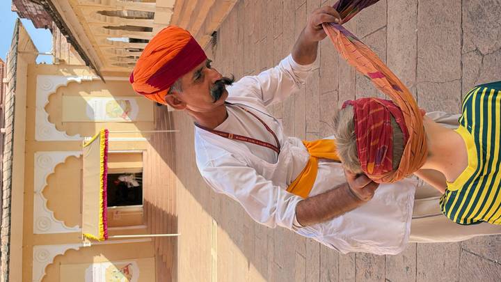 Turbaned man carefully wraps a colorful turban on a young boy in a sunlit courtyard of a historic Indian palace.
