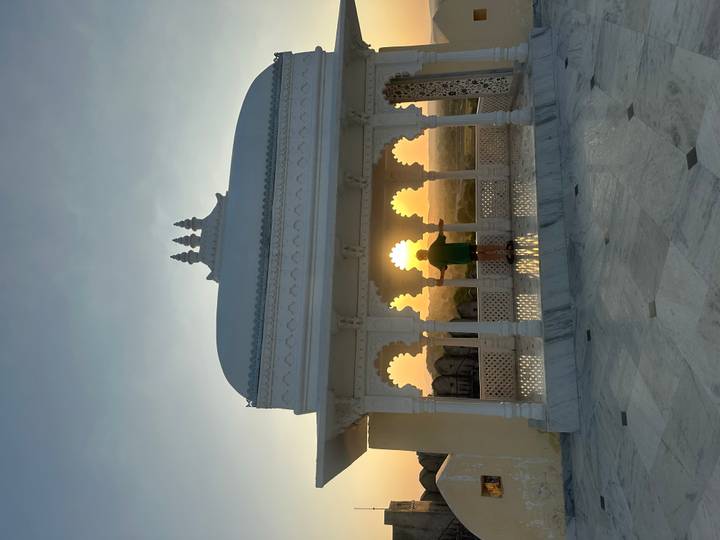 Silhouette of a person framed by ornate arches catching the setting sun from a white marble pavilion.