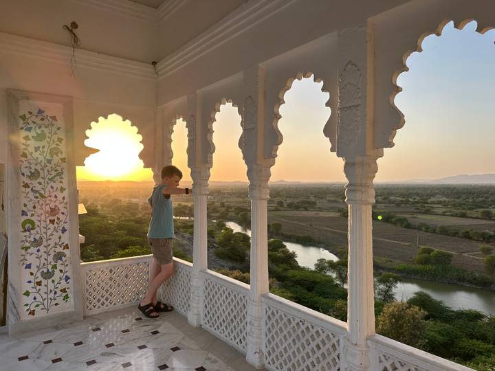 Boy leans on a white lattice balcony enjoying a golden sunset over a winding river and green countryside.