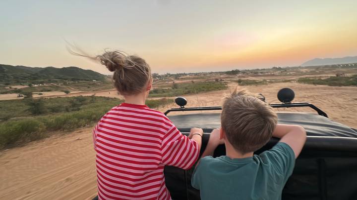 Two children ride standing in an open jeep across sandy terrain at sunset with hair blowing in the wind.