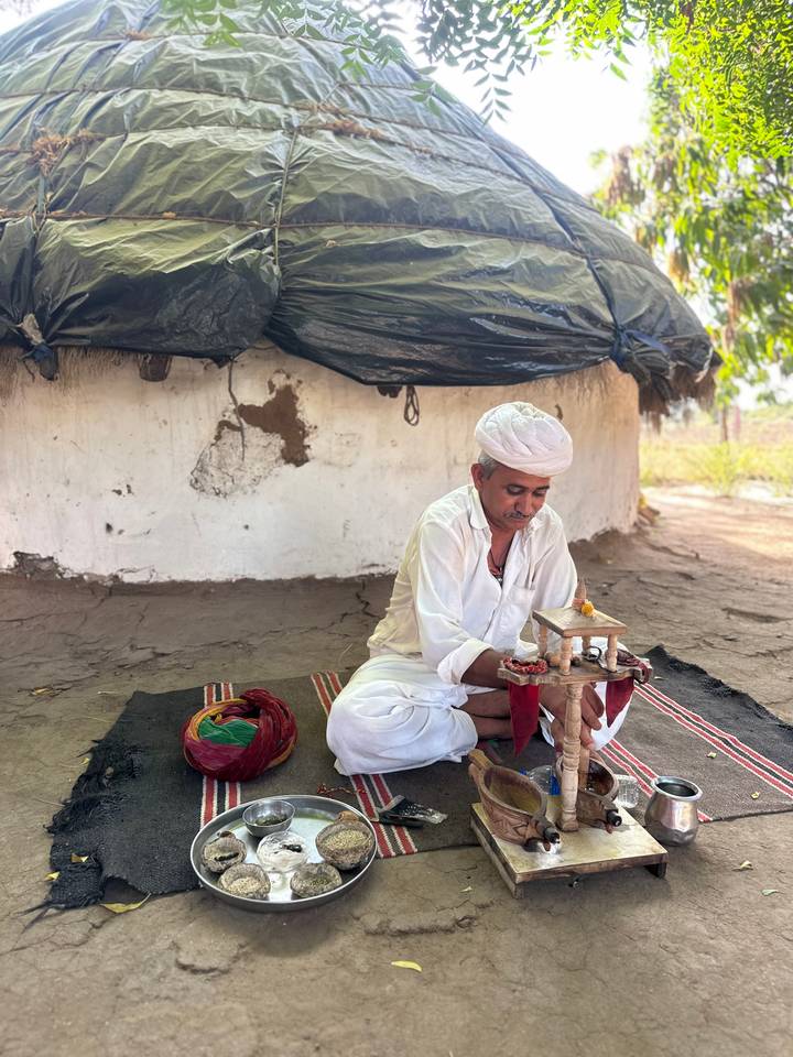 Elderly man in white traditional attire performs a ritual seated on a mat outside a mud hut.