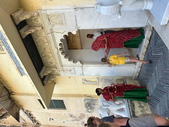 Women in red shawls greet a young girl at a carved marble doorway offering a ceremonial tray.