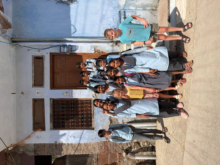 Group of local schoolchildren pose smiling with two visiting kids outside a blue-painted building.