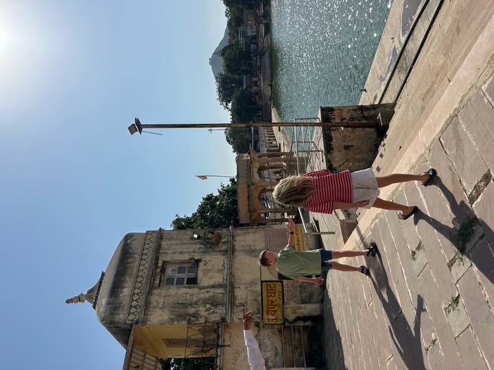 Two children wander along the stone steps of a lakeside ghat beside turquoise water under a bright sky.