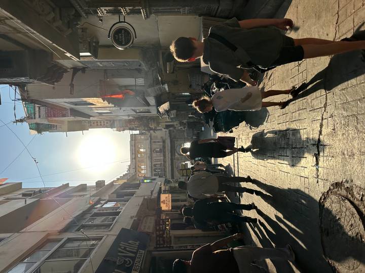 Backlit street scene with tourists and locals walking through a narrow alley of shops in the late afternoon sun.