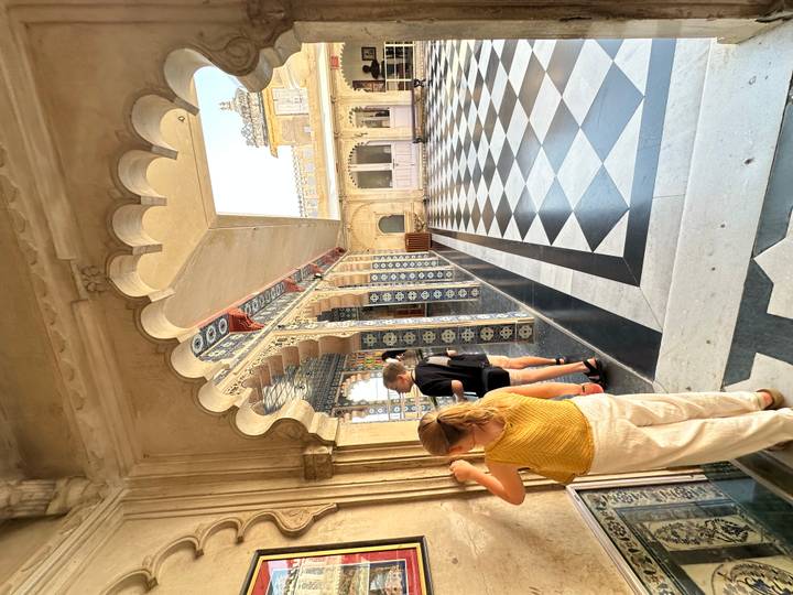 Tourists examine ornate blue-tiled arches and checkerboard floors inside a palace corridor.