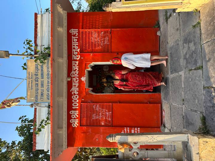 Two visitors enter a bright red Hanuman temple with Hindi script and decorative bells above the doorway.