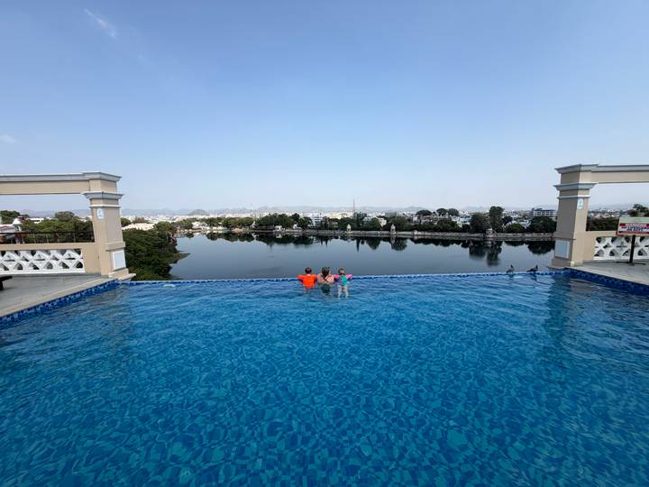Two children enjoy an infinity rooftop pool overlooking a tranquil lake and cityscape on a bright day.