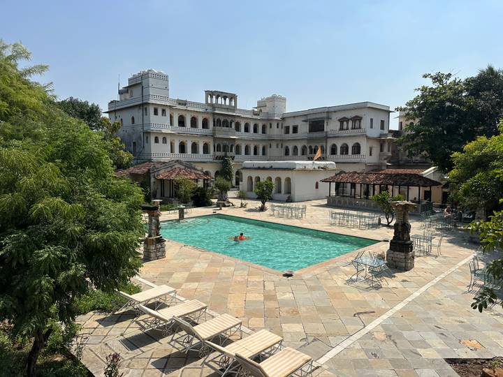 A heritage palace hotel surrounds a turquoise courtyard pool where a lone swimmer floats.