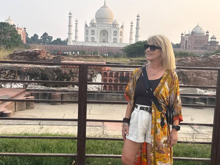 A woman standing near ruins with the Taj Mahal visible across the lawn in the background.