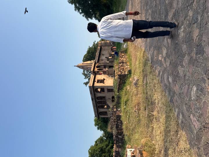A man walks toward a rustic stone temple building set in dry grassland beneath a clear sky.