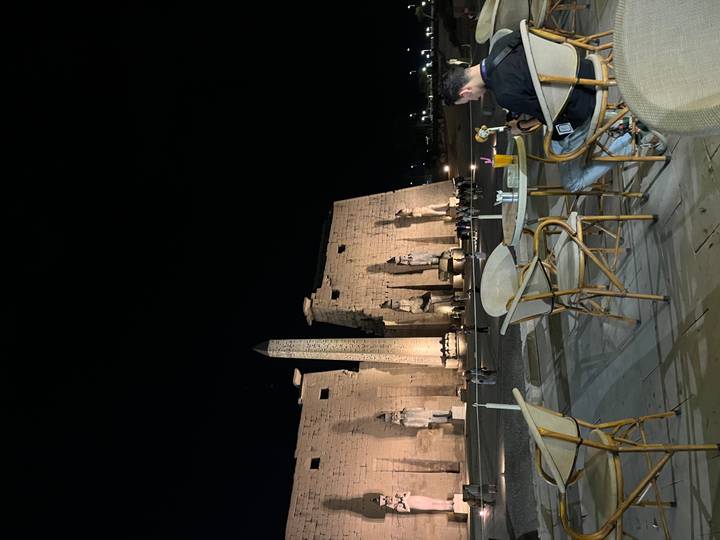 Outdoor café tables with Luxor Temple obelisk and statues dramatically lit at night in the background.