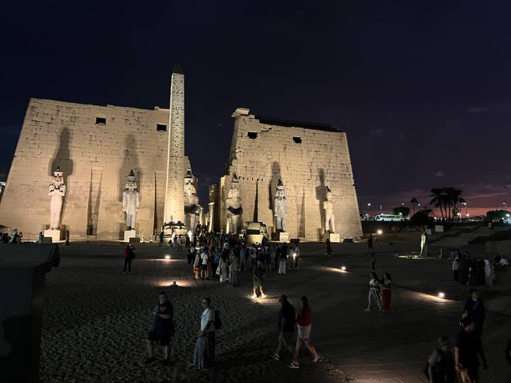 Crowd gathers at night inside Luxor Temple complex illuminated by warm lights with purple dusk sky.