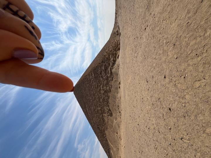 Playful perspective shot of a hand appearing to pinch the top of a small pyramid against a wispy sky.