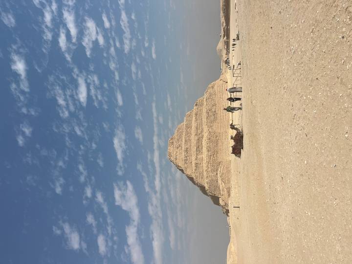 The Step Pyramid rises from the desert sands with a camel caravan and a few visitors in the foreground under a blue sky with scattered clouds.