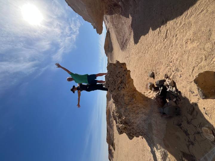 Two travellers stand triumphantly on a weather-carved rock outcrop in a wide desert landscape beneath a bright blue sky.