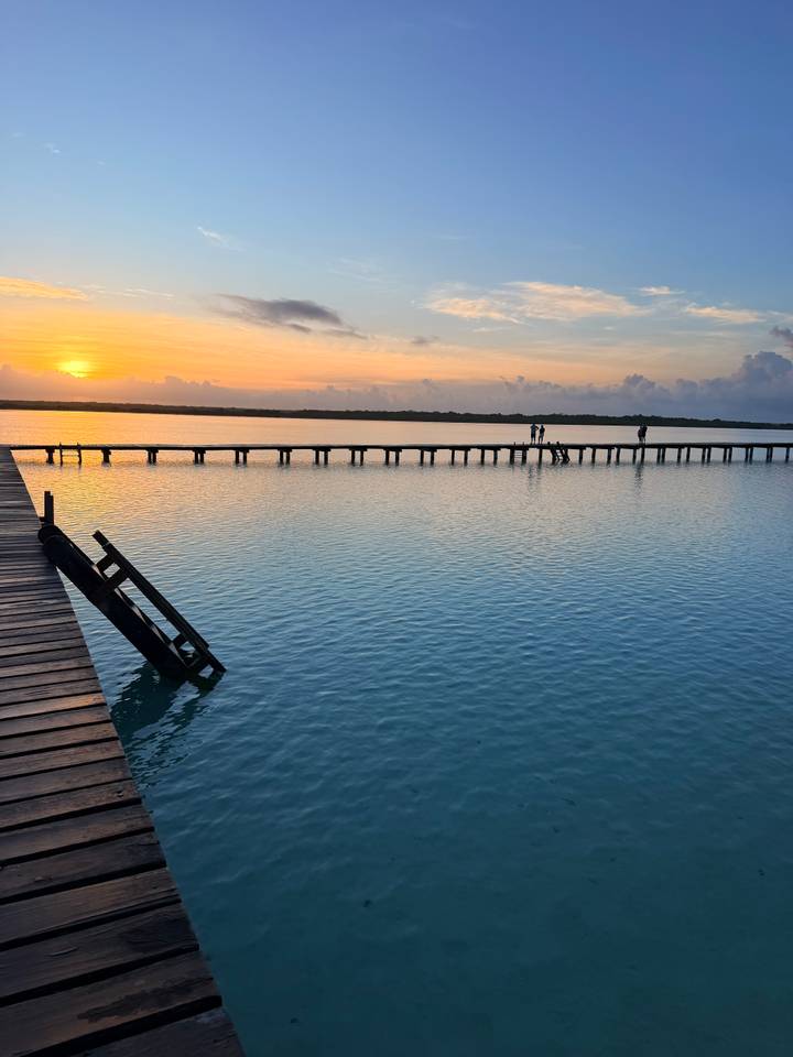 A calm lagoon reflects warm sunset hues while a wooden pier stretches toward the horizon with tiny silhouettes of people.