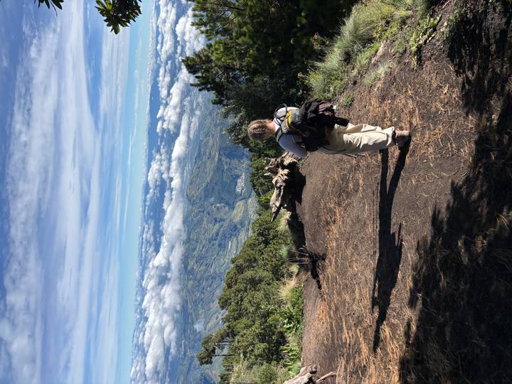A trekker with a backpack walks along a volcanic ridge with sweeping cloud-filled valleys below.