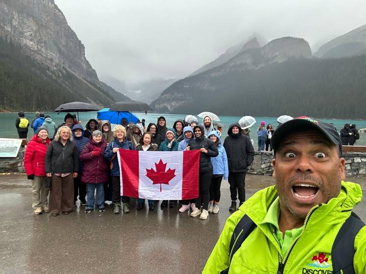 A tour group holds a Canadian flag on a rainy day beside the turquoise waters and mountains of Lake Louise.