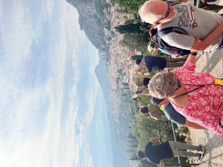 Tour group taking photos over Taormina rooftops toward hazy Mount Etna.