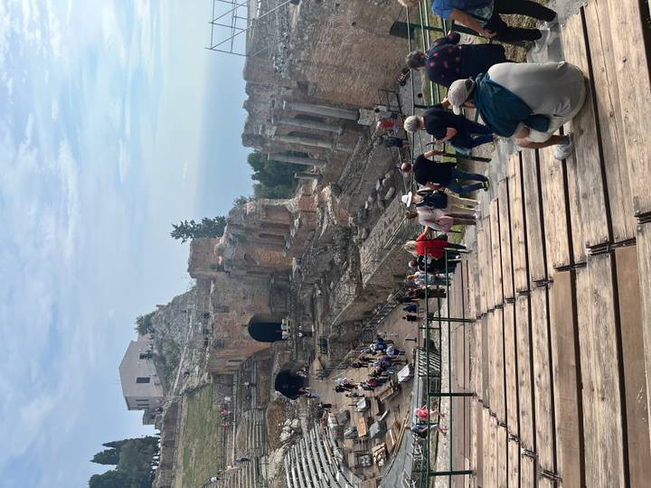 View down wooden seating rows into Taormina’s ancient Greek theatre with visitors exploring the stage area.