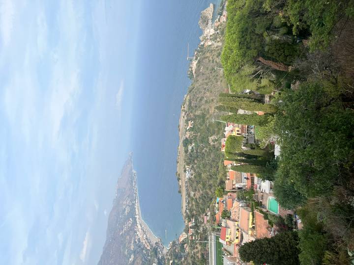 High coastal panorama over Taormina and the turquoise Ionian Sea with cypress trees in foreground.
