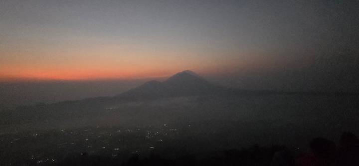 Pre-dawn silhouette of a volcanic peak against a faint orange horizon.