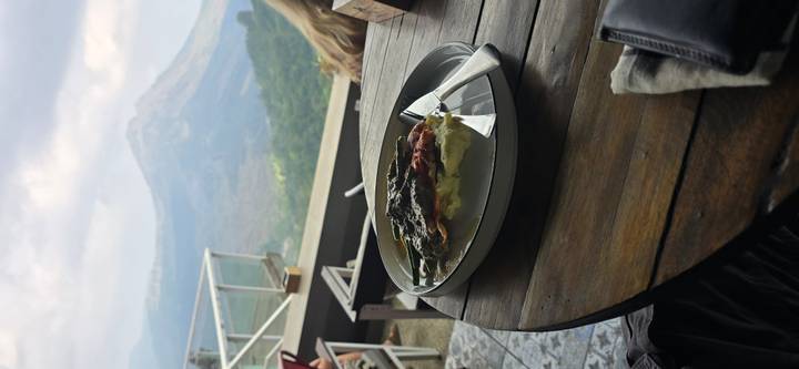 A plated meal on a café table overlooks a hazy volcanic landscape through glass railings.