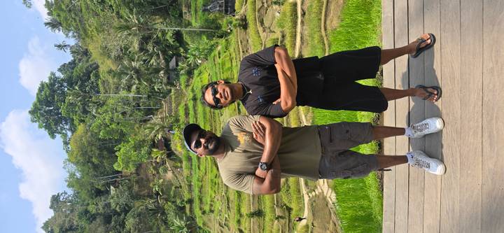 Two men pose confidently in front of terraced rice fields and tropical forest.