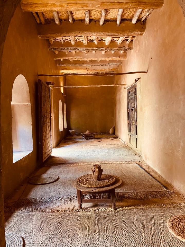 Simple earthen interior room with arched doorways, woven mats and rustic furnishings lit by soft window light.