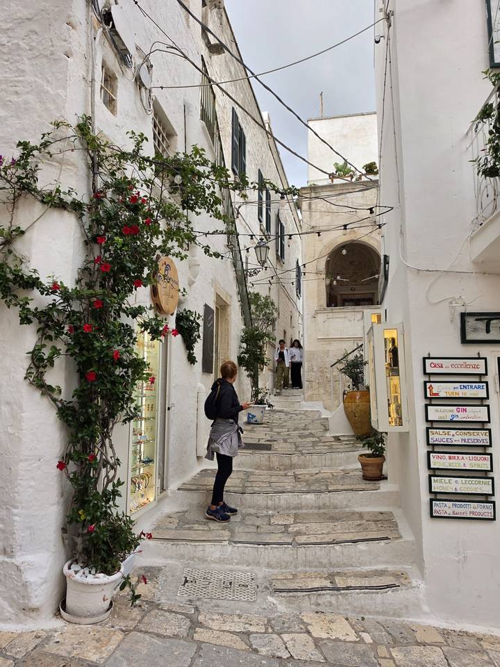Narrow stone alley lined with flowers and small shops as a woman takes a photo.