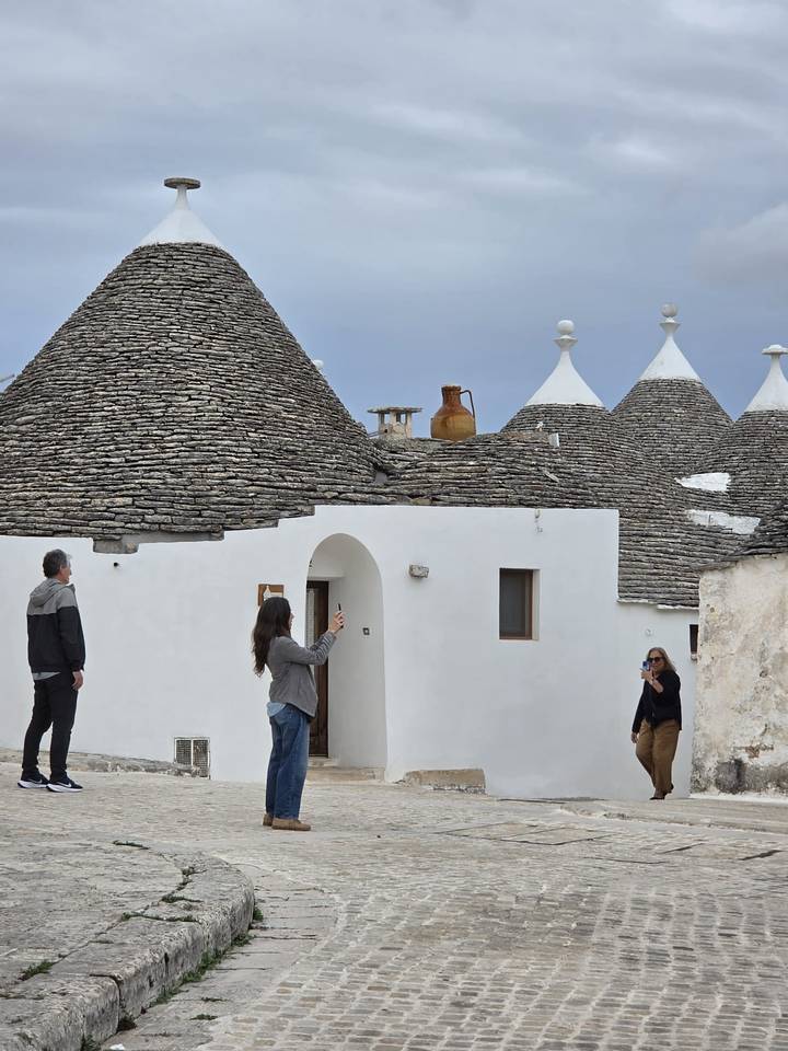 Visitors photograph the whitewashed trulli houses with conical stone roofs in a small lane.