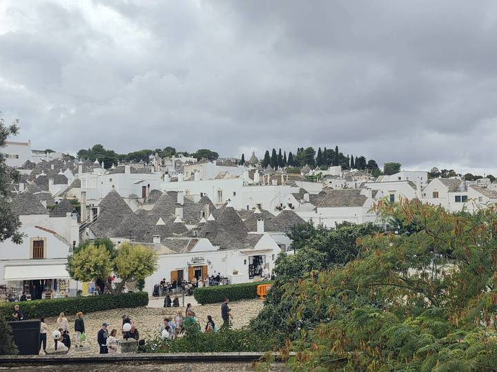 Panoramic view over a hillside town filled with dozens of grey-roofed trulli under a cloudy sky.