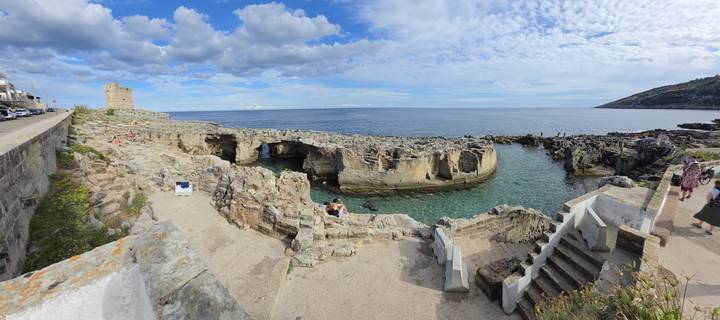 Rocky coastal inlet with clear turquoise water, stone steps and a medieval watchtower under a patchy sky.