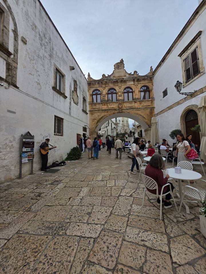 Busy cobbled lane with café tables, an archway and a street musician playing guitar.