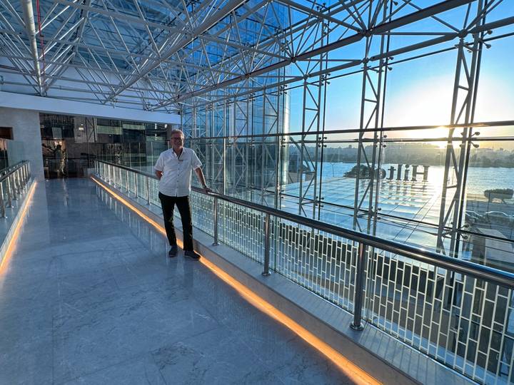Man stands on glass-roofed walkway overlooking the Nile at sunset inside modern terminal.