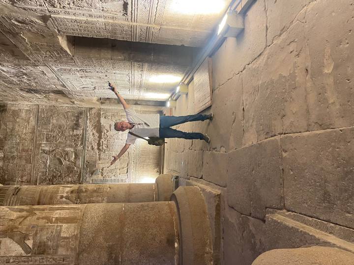 Visitor gestures triumphantly in sandstone temple hall adorned with hieroglyphic reliefs.