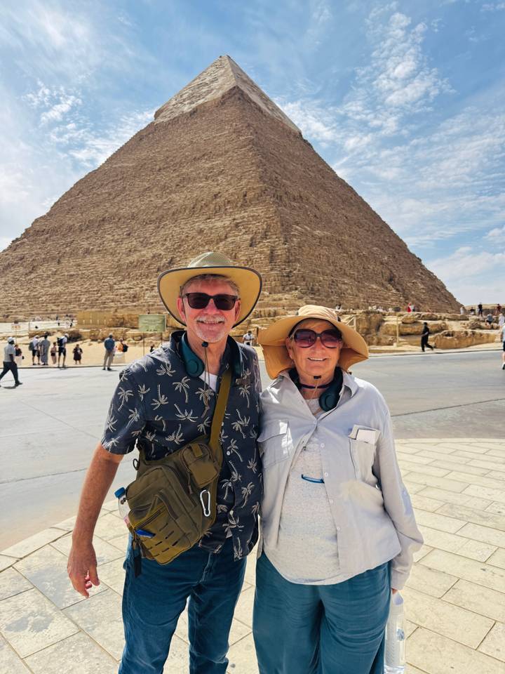 Two travellers in sun hats smile with Great Pyramid rising behind them on Giza Plateau.