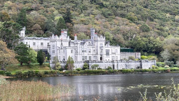 Kylemore Abbey’s grey stone façade reflected in a calm lake at the foot of a forested hillside.