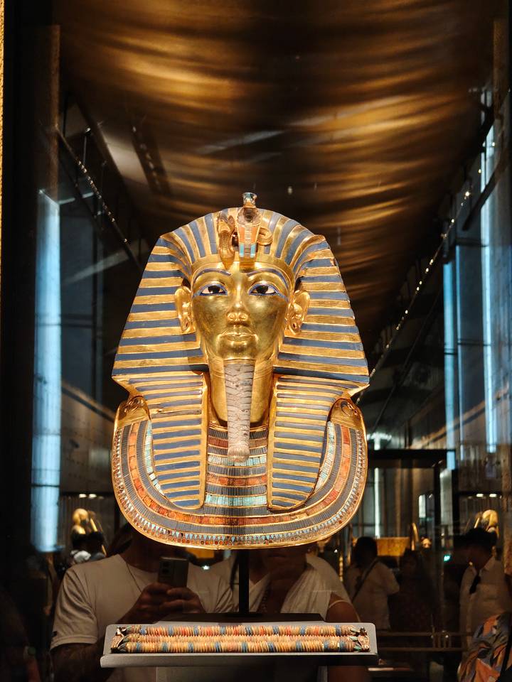 Gold funerary mask of Tutankhamun displayed under spotlights inside a museum gallery.