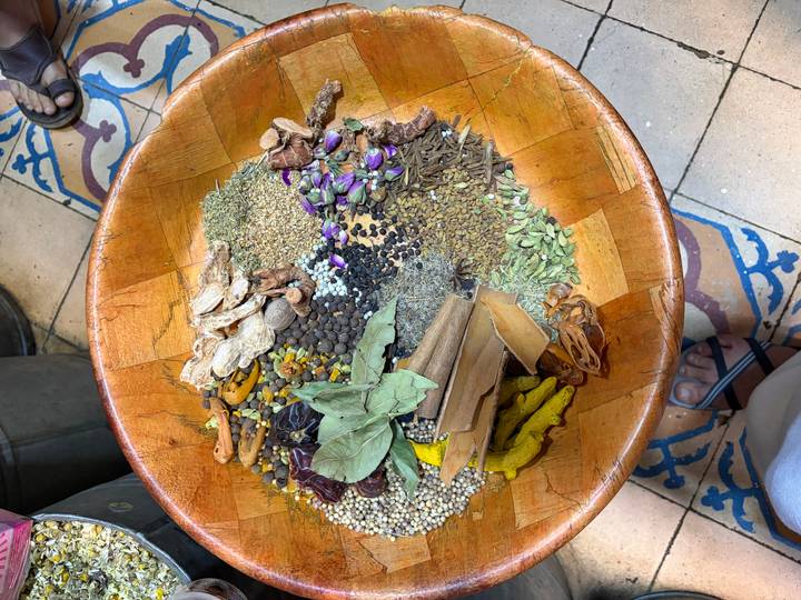 Large wooden bowl filled with assorted spices, herbs and dried ingredients held over a tiled floor.