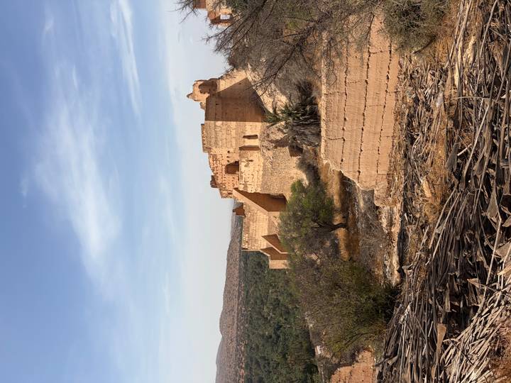 Ruined desert kasbah standing on a hill above sparse vegetation and dry terrain.