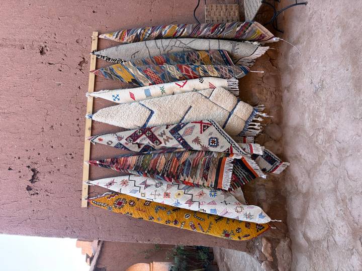Row of colorful hand-woven Moroccan rugs hanging against an earthen wall.
